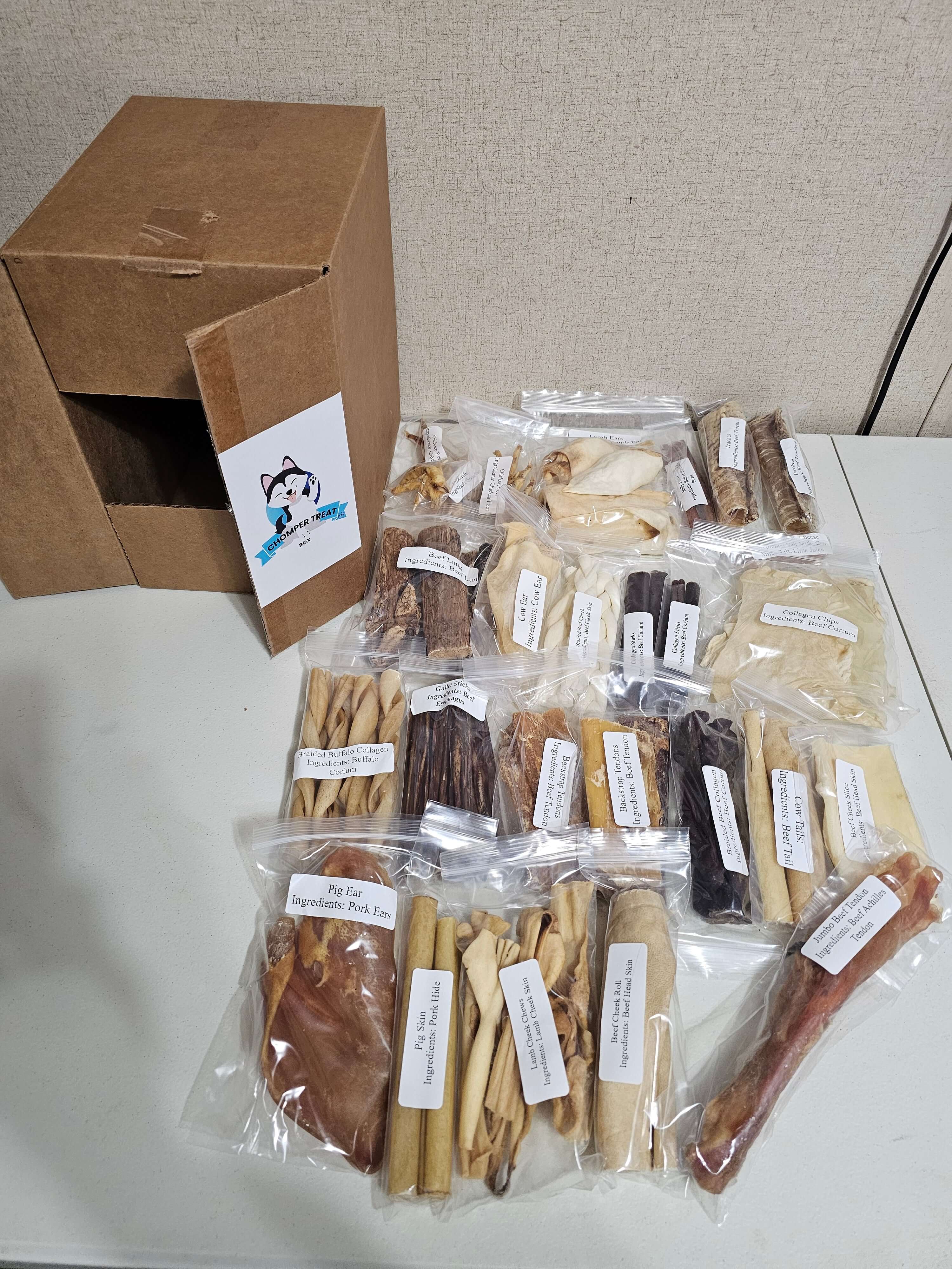 Deluxe Chomper Treat Box with 60 all-natural dog chews in clear bags, arranged beside a cardboard box.